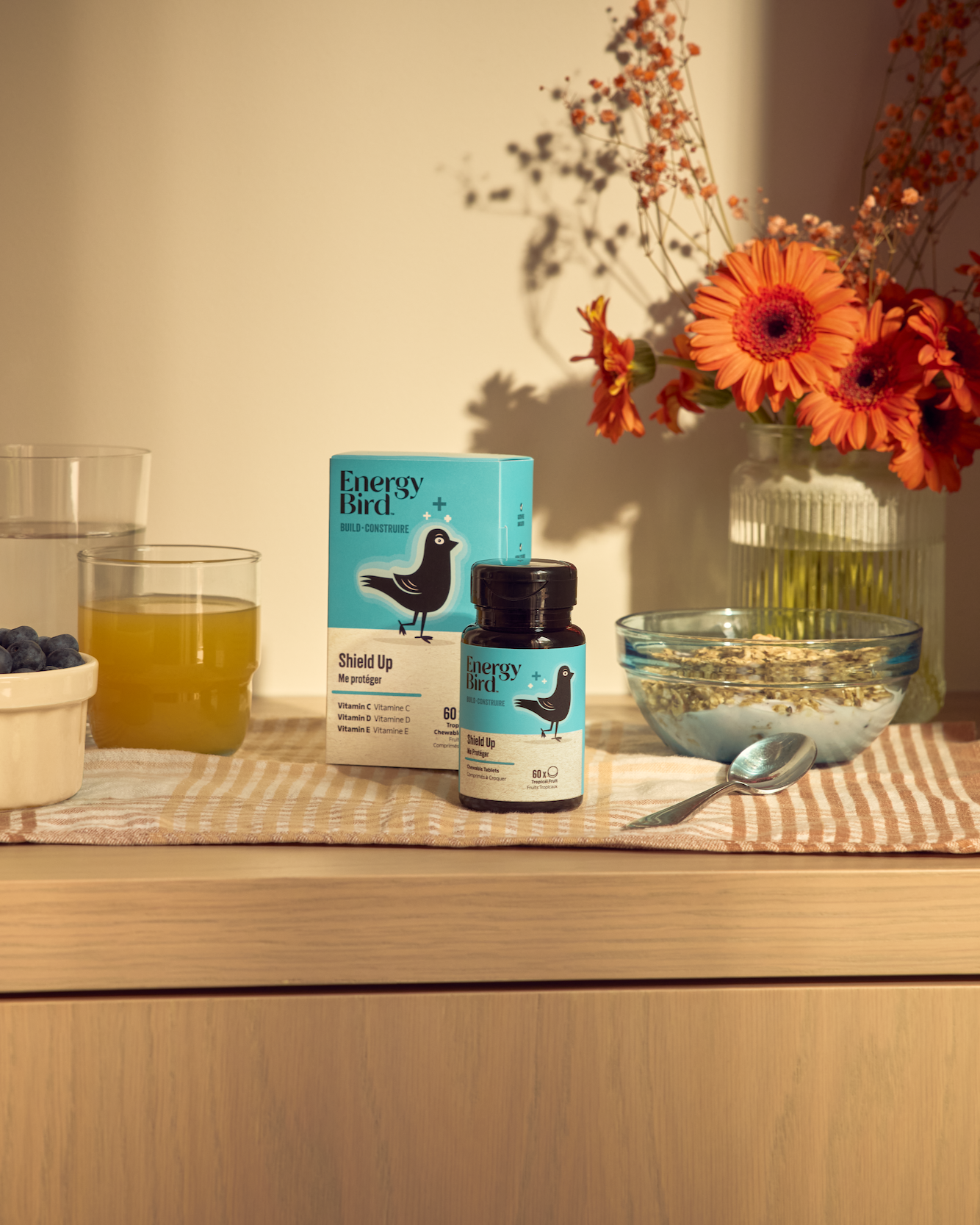 Energy Bird's Shield Up Packaging and Bottle on a dining table beside a bowl of cereal, bowl of blueberries and a cup of orange juice